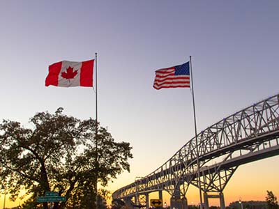A Canadian and American flag side by side waving in the wind in front of the Sarnia bridge.