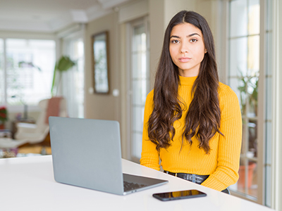 Young woman sitting at a kitchen counter with a laptop and phone looking into the camera with a neutral expression