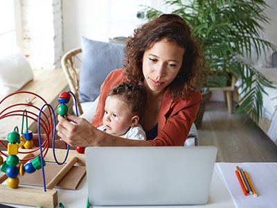 A mother working from home on a laptop with her baby on her lap, distracting the baby with a toy.