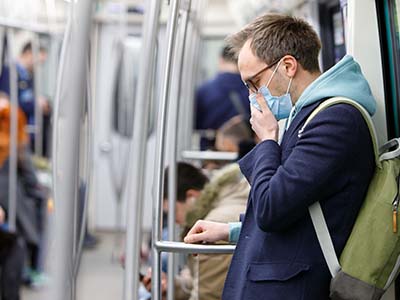 Man on a bus, wearing a blue surgical mask, holding his face like he's coughing.