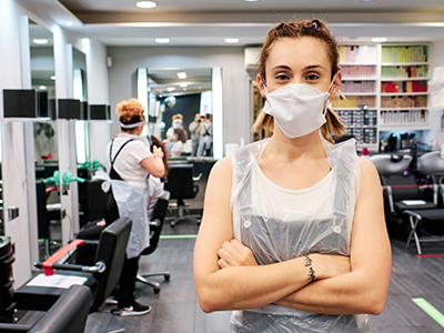 Hair stylist standing in a salon looking into camera with crossed arms, wearing a mask and other PPE