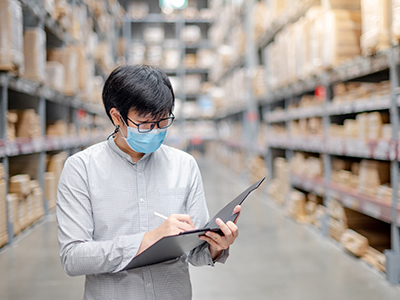 Man in a warehouse, looking at a clipboard while wearing a mask.