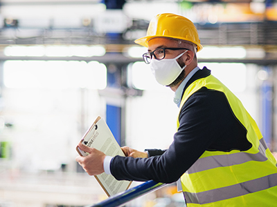 Man with glasses wearing a white mask, yellow hard hat and safety vest, leaning on a railing and holding a clip board.