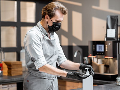 A man standing behind a counter in an apron, wearing gloves and a face mask, sanitizing hands.