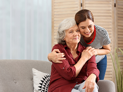 Older woman sitting on couch while younger woman stands and hugs older woman from behind.