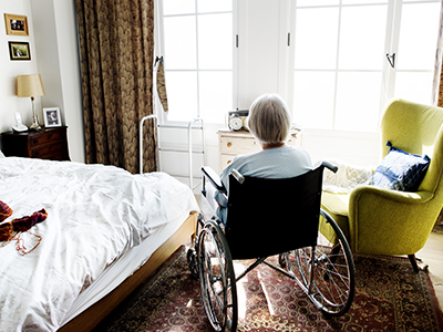 Older woman with her back to the camera, sitting in a wheel chair in her bedroom looking out the window.