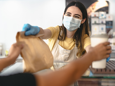 Female worker wearing a mask and gloves handing coffee and a bag to a customer.