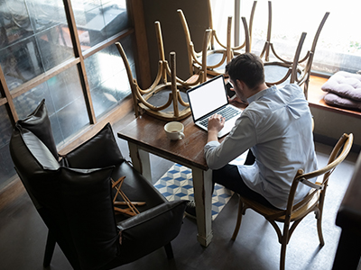 Aerial view of restaurant owner sitting at a table working on his laptop. The additional chairs are turned over onto the table.