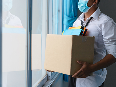 A man wearing a blue surgical mask holds a box of desk items. He is looking out of a office window.