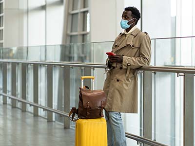Man wearing a mask standing in an airport with his yellow roller suitcase and brown backpack