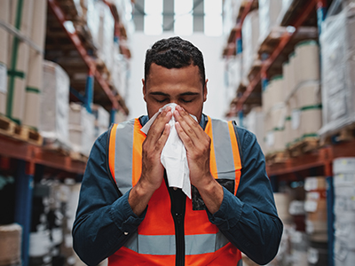 A warehouse worker in an orange vest, standing between warehouse shelves, blowing his nose with a tissue.